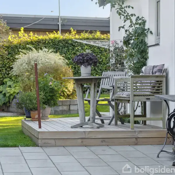 Terrasse med træbord og bænk med hynder. Bordet har en plante på toppen. Omkring terrassen er der grønne buske og træer, der skaber en hyggelig haveatmosfære.
