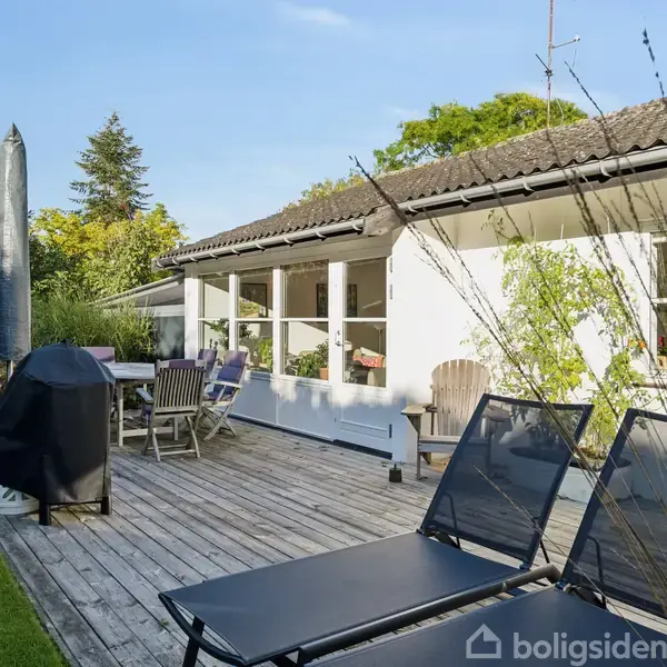 En træterrasse med liggestole og en grill, omgivet af en grøn have. Et hvidt hus med store vinduer i baggrunden.