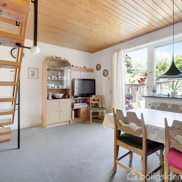 Rektangulært spisebord med stole rundt står i en stue med trægulv og lysindfald fra store vinduer. Trappe til venstre, reol med porcelæn og et fjernsyn i baggrunden.