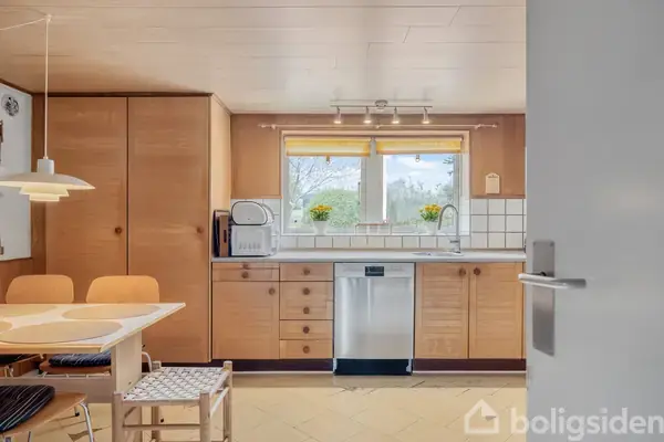 Køkken med træskabe og bordplade, indeholder opvaskemaskine og mikroovn. Bord med stole og lampe hængende over. Vindue med udsigt til træer, omgivet af fliser med to potteplanter på kanten.