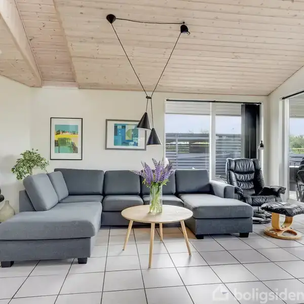 Grå sofa i stue med lyse fliser, flankeret af sorte lænestole. Over sofaen hænger to lamper. Væggene har farverige billeder, mens store vinduer fører ud til en terrasse.