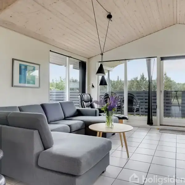 Grå sofa står i en lys stue, dekoreret med moderne kunst på væggene. Store vinduer slipper naturligt lys ind, mens en glasdør åbner mod en træbalkon.
