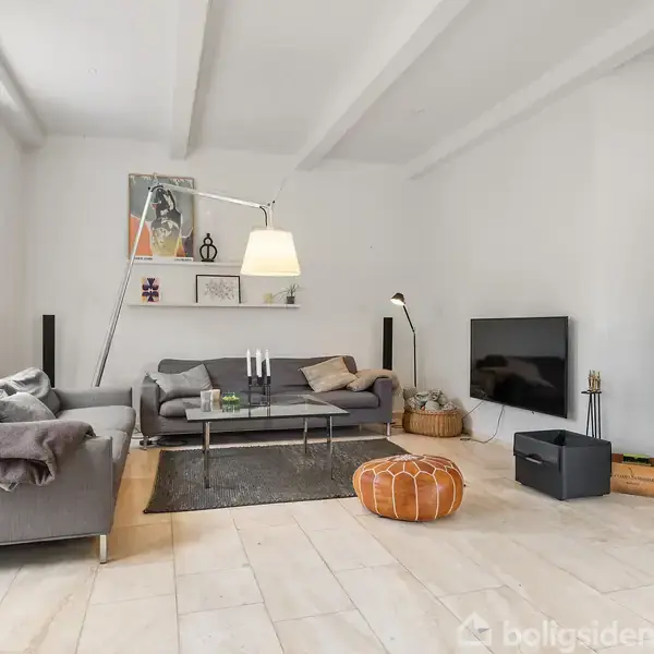 En stue med grå sofaer og et sofabord står på et lyst trægulv. En moderne brændeovn i hjørnet, og en fladskærms-TV ved siden af. Lys og æstetisk indretning.
