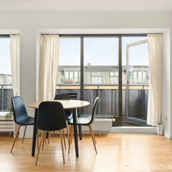 Et rundt spisebord med sorte stole står i et lyst rum med trægulv. Store vinduer med hvide gardiner åbner ud mod en balkon med udsigt til tage.