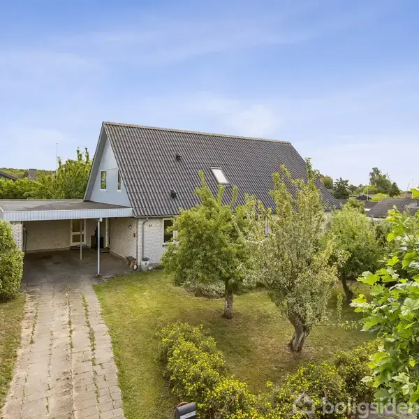 Et hvidt hus med skråt tag står for enden af en flisebelagt indkørsel. Tæt omkring er der en velplejet have med træer og buske under en blå himmel.