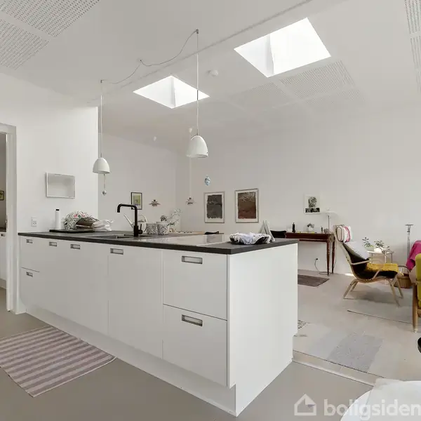 Køkkenet har en stor hvid køkkenø med sorte bordplader. Der er hængelamper over øen og ovenlys i loftet. Stuen i baggrunden har farverige møbler og vægdekorationer.