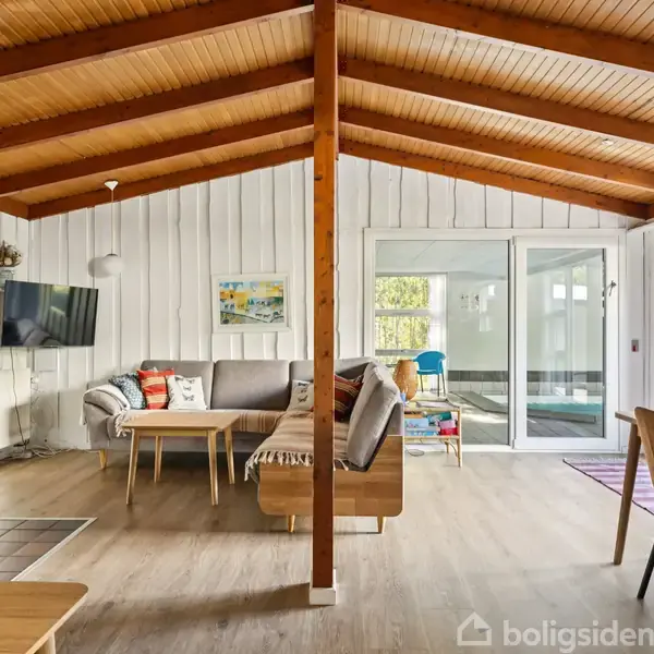Sort træhus med lavt tag står i en grøn have. Omkring huset ses en skov med tætte træer, der skaber en fredelig og naturlig atmosfære.