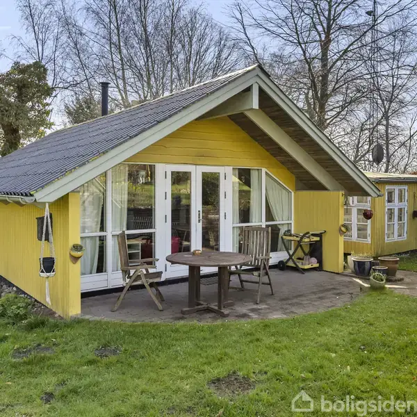 Et gult sommerhus står på en græsplæne med en terrasse foran. På terrassen er der et bord og stole. Baggrunden har træer uden blade.