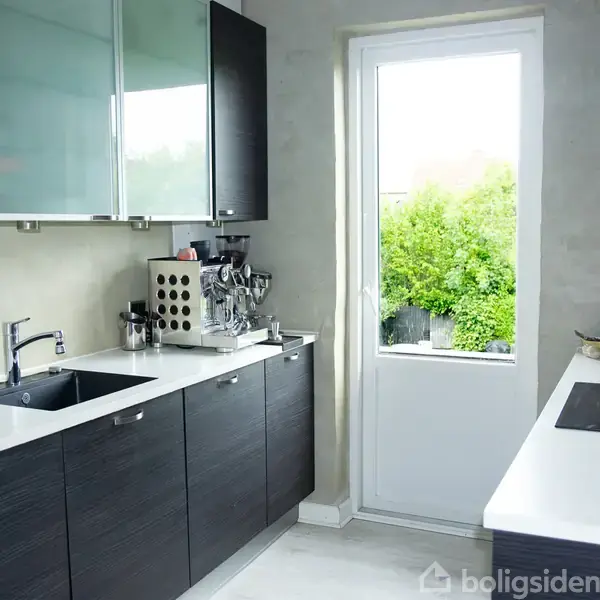 Et moderne køkken med sorte skabe og hvid bordplade er i brug. En kaffemaskine står ved siden af vasken. Gennem et vindue ses en grøn have.