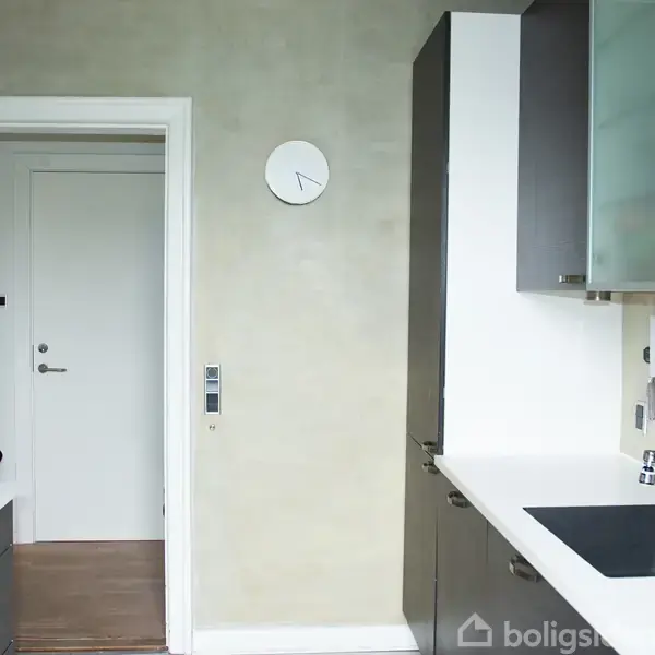 Ur på væggen viser tiden; køkkenvask med hane er til højre; køkkenskabe og skuffer omgiver området; en dør fører til et tilstødende rum i baggrunden.