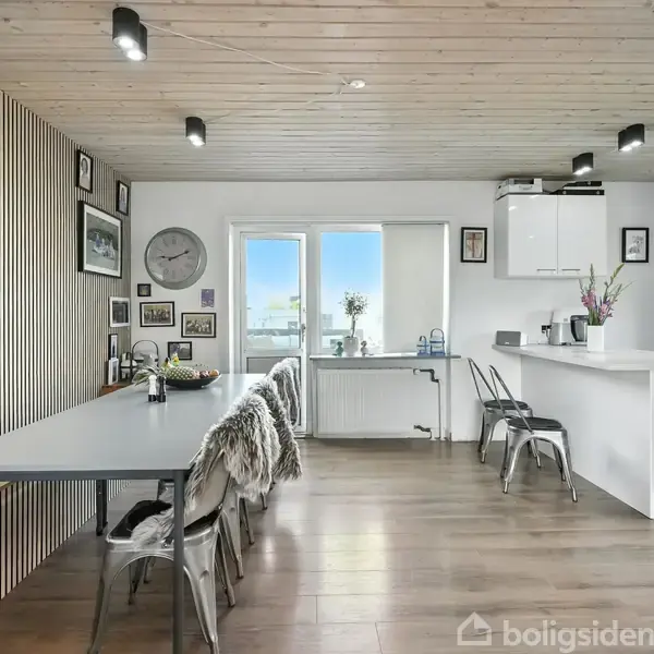 Et moderne køkken-alrum med et langt spisebord på venstre side, dekoreret med stole og bløde tæpper. Til højre findes en hvid køkkenø med barstole. Store vinduer giver naturligt lys. Der er ingen tekst.
