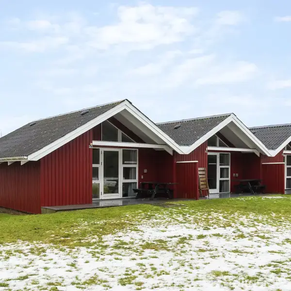 Tre røde træhytter står i række på en græsplæne med nogle pletter af sne. De har store vinduer og gavltage, skaber et hyggeligt udtryk i et åbent område.