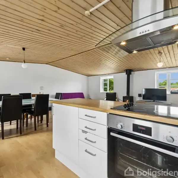 Køkken med hvidt skab og indbygget ovn, i et åbent rum. Et spisebord og sorte stole står ved siden af, med træloft og et vindue i baggrunden.