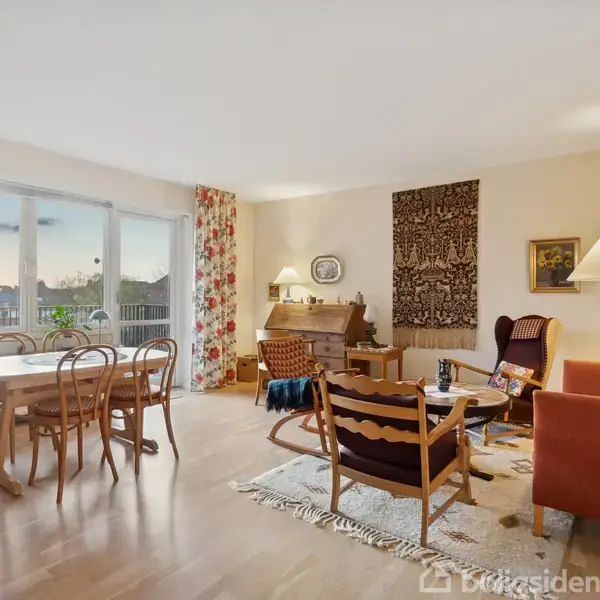 Stue med spisebord og stole ved vindue, sofa og lænestole omkring sofabord, omgivet af tæpper og lamper. Store vinduer giver udsigt til solnedgang. Farverige gardiner tilføjer liv.