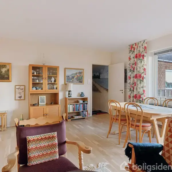 Stue med træmøbler og sofa, dekoreret med malerier og en bogreol, samt et spisebord med stole nær et vindue med udsigt til en murstensbygning.