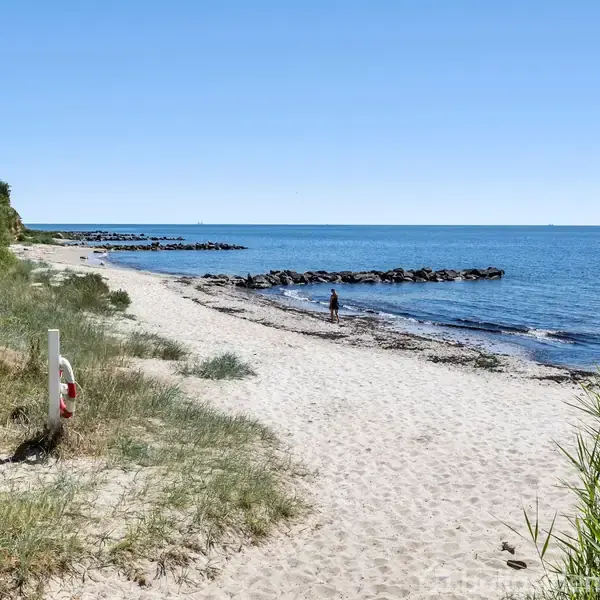 Strand med hvidt sand strækker sig langs havet. En person går ved vandkanten. Grønt buskads og klipper ses langs strandens kant under en klar blå himmel.