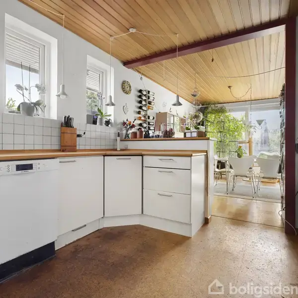 Køkkenet har hvide skabe og træbordplader, mens planter står på bordet. I baggrunden ses et spiseområde med glasdør ud til en terrasse, omgivet af grønt og naturligt lys.