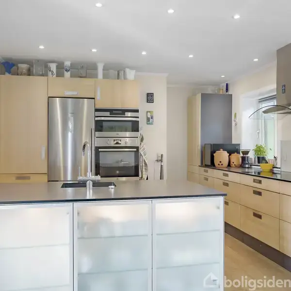 Et moderne køkken med en stor køkkenø i forgrunden. Bagved ses indbyggede træskabe, en stålfarvet køleskab, ovn og mikroovn. Bordpladen har krydderier og urter. Lys strømmer ind fra et vindue.