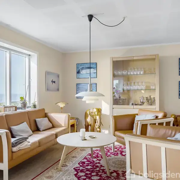 En stue med lysebrune sofaer omkring et rundt bord, og en loftslampe over det. Store vinduer giver udsigt til naturen. Vægge er dekoreret med billeder og reoler med glas.