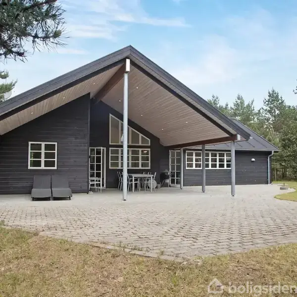 Et sort sommerhus med spidst tag står på en flisebelagt terrasse. Der er solstole og et bord- og sæt under overdækket. Omgivet af grønne træer og buske.