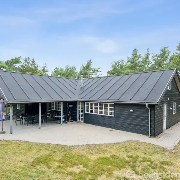 Sort træhus med gråt tag står på en stor terrasse. Der er en overdækket veranda med havemøbler. Omgivet af græs og fyrretræer under en klar blå himmel.