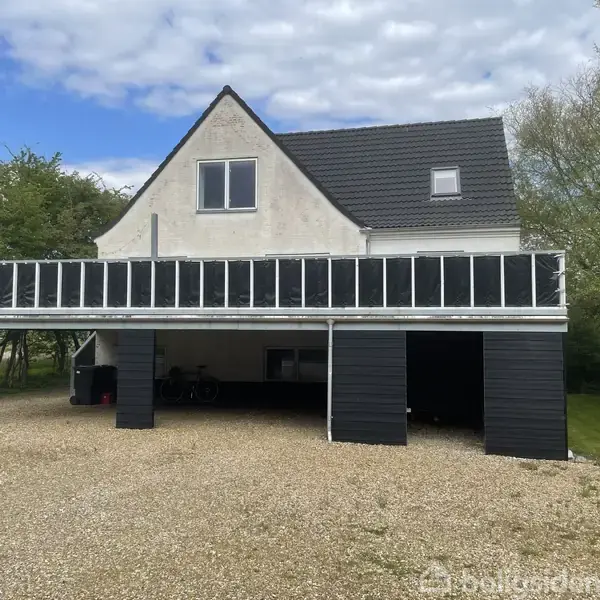 Hvidt hus med sort tag har hævet terrasse med glasrækværk. Under terrassen er der carport med grusbelægning. Omgivet af træer og græsplæne. Skyfri himmel med spredte skyer.