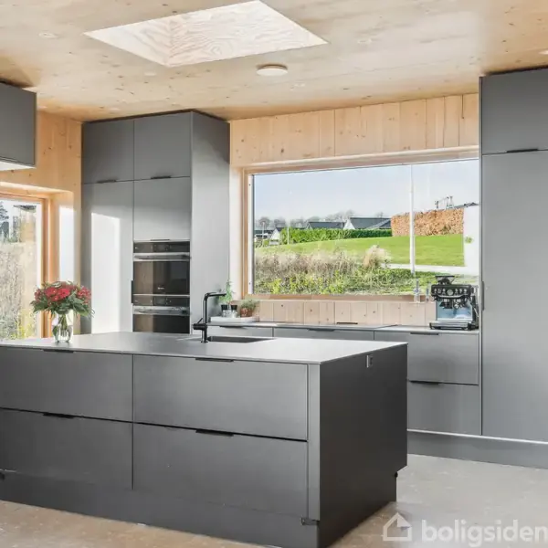 Moderne køkken med stor, mørk køkkenø, omgiven af grå skabe. En bordplade med frisk blomsterdekoration og kaffemaskine ved et stort vindue med udsigt til grønne områder.