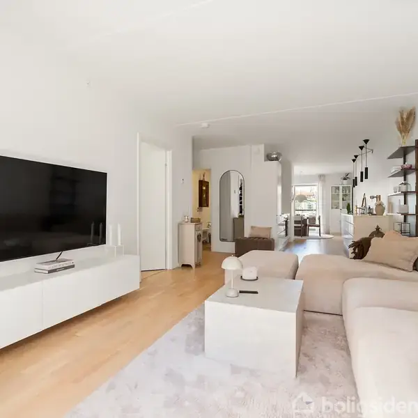 En moderne stue med en stor fladskærms-TV på en hvid skænk. En lys sofa står mod en væg, mens en hylde og kunst dekorerer rummet bagved.