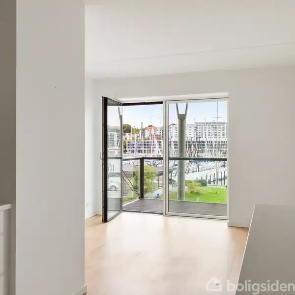 En lys stue med en stor fladskærms-TV placeret på et hvidt mediebord. En lys sofa og et sofabord står i rummet, der har trægulve og åbner op til et køkkenområde.