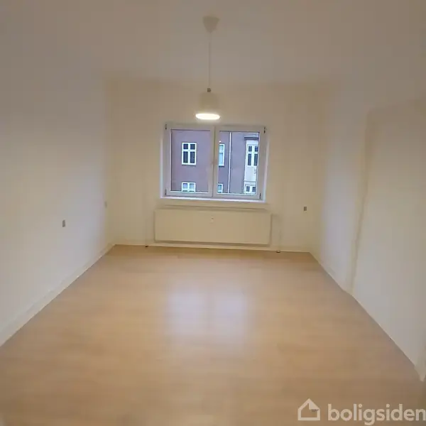 Et tomt rum med trægulv og hvidmalede vægge indeholder en enkel loftlampe, der hænger centralt. Et stort vindue viser en udsigt til en murstensbygning udenfor.