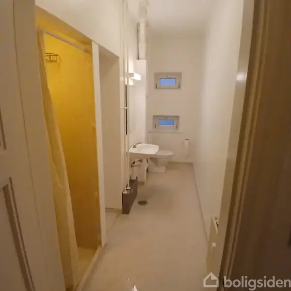 Badeværelse med hvid håndvask og toilet. Til venstre en bruser med gul flisevæg. Væggene er lyse, og der er to små vinduer over toilettet. Gulvet er lyst.