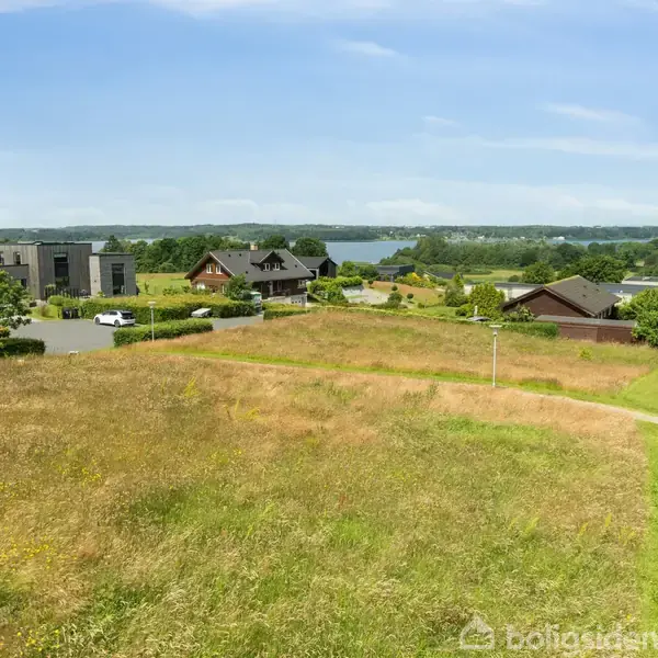 Græsareal strækker sig over landskab med moderne huse i baggrunden. En vej leder mod huse, og en sø kan ses i horisonten under en blå himmel.