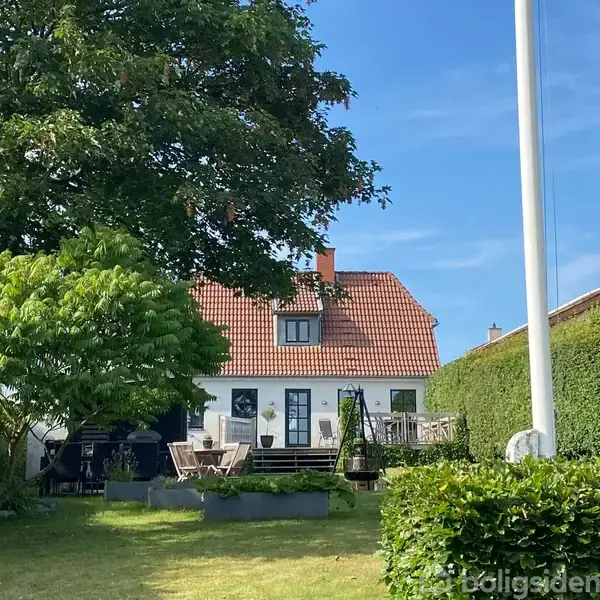 Et rødt tegltagshus med terrasse står omgivet af en frodig have. Træer og hækkeklippede buske omkranser huset, mens en flagstang står i forgrunden under en blå himmel.