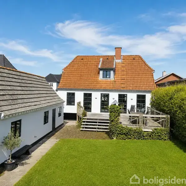 Et hvidt hus med rødt tegltag står i en velholdt have. Træterrasse foran indgang, flankeret af grønne buske. Blå himmel og andre huse ses i baggrunden.