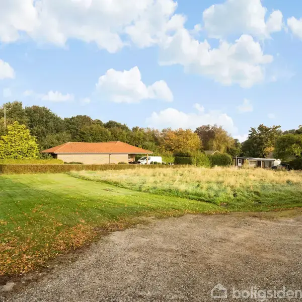 En grusvej fører til et hus med rødt tag omgivet af en stor, grøn græsplæne og buske, placeret i et naturskønt område med træer under en klar blå himmel.