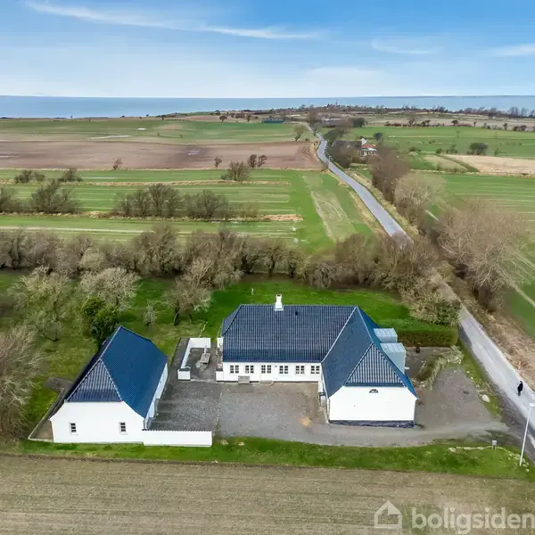 Hvidt hus med blåt tag ligger i et landligt område omgivet af marker og en vej. I baggrunden ses kysten og havet.
