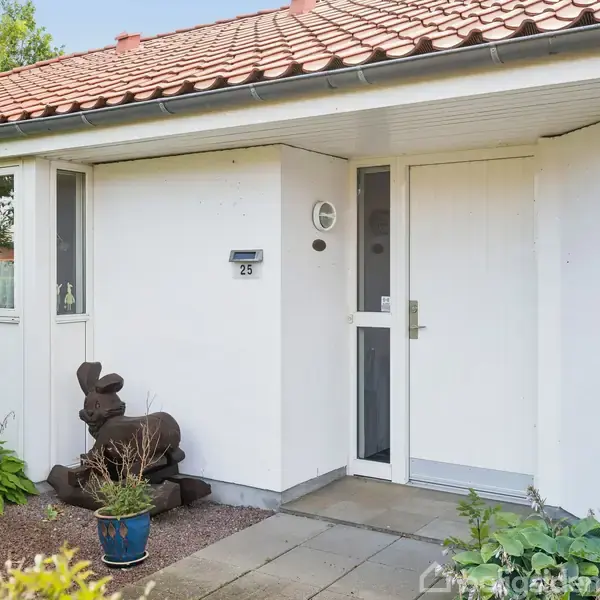 Hvid husfacade med en brun skulptur ved siden af indgangsdøren. Nummer 25 står på en postkasse. Omkranset af grønne planter og flisebelagt sti.