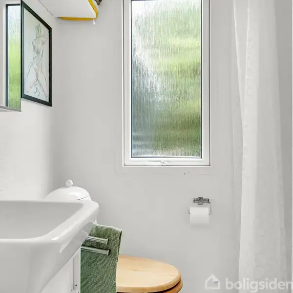 Håndvask med skab og spejl over, står i et lille badeværelse med vindue i baggrunden. Hvid bruserforhæng til højre, toiletpapirholder under vindue, og kunstbillede på væggen.
