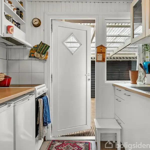 Køkken med hvid indretning og køkkenudstyr, dør står lidt åben. Hylder rummer krydderier, og håndklæder hænger ved ovn. Farvestrålende køkkenting hænger fra kroge ved vask. Klokke på væggen.