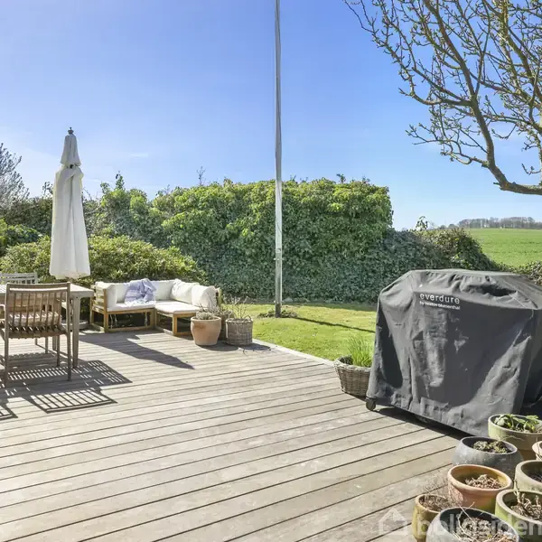 En træterrasse med havemøbler og en grill under et betræk. Potteplanter står langs kanten. Baghaven grænser op til en hæk med udsigt over et grønt landskab.