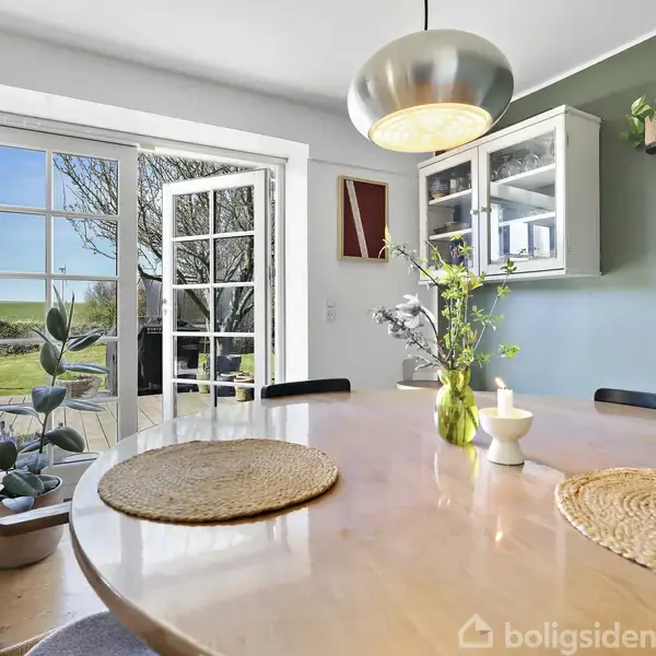 Rundt spisebord med flettede dækkeservietter og blomster i vase. Store, åbne glasdøre slipper lys ind. Køkkenområde med skab og planter på væggen. Grøn have ses udenfor.