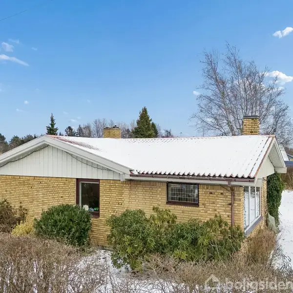 Et murstenshus med saddeltag dækket af sne ligger i en have med buske. Ved siden af huset står en grå bil. Himmelen er blå med få skyer.