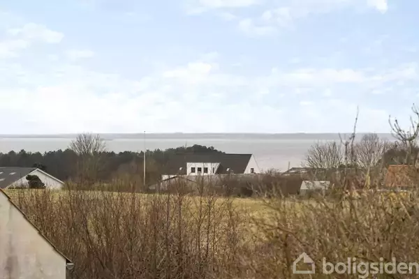 Huse står stille blandt træer og buske i et åbent landskab med havet i baggrunden under en overskyet himmel.