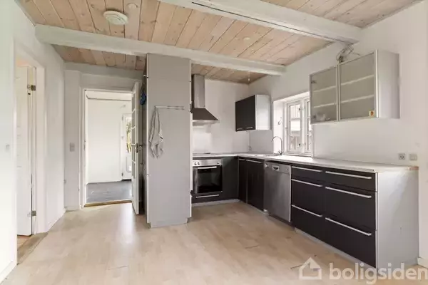 Et køkken med mørke skabe og stålgreb, åbne hylder, en indbygget ovn og en emhætte. Lyse trægulve og lofter skaber en lys og rummelig atmosfære.