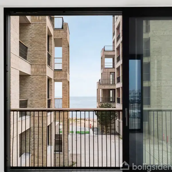 Udsigt fra en moderne lejlighed med store vinduer ud mod gaden. To murstensbygninger flankerer siden, mens havet er synligt i det fjerne mellem bygningerne.