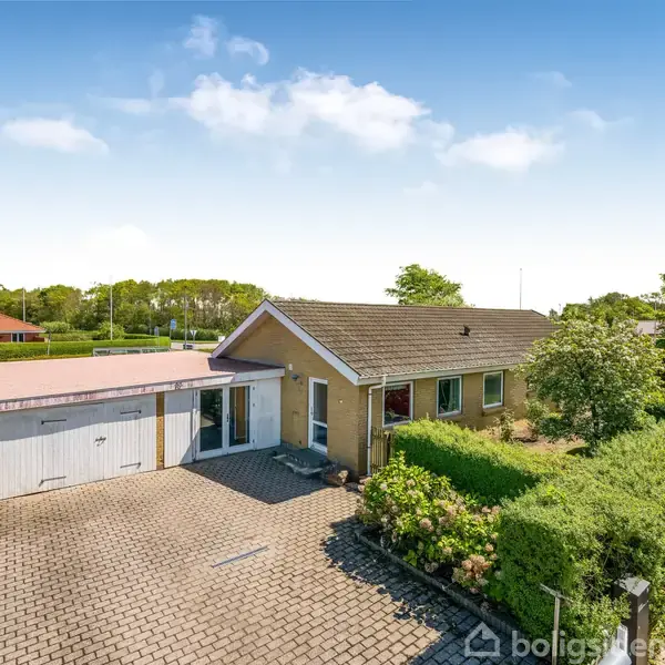 Et enkelt hus med gavltag og garage står ved siden af en brostensbelagt indkørsel. Omkring huset er der grønne buske og træer i en boligområde.