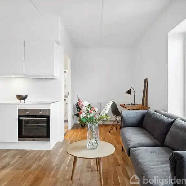 Sofa står i moderne stue-køkken, ved siden af et rundt sofabord med blomster. Køkkenet har hvide skabe og en indbygget ovn. En arbejdsplads med lampe ses i baggrunden.
