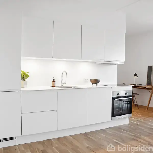 Et moderne køkken med hvide skabe og en indbygget ovn står i et lyst rum. På bordet ses en plante og håndsæbe. Ved siden af er et arbejdsområde med skrivebord og stol.