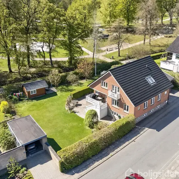 Et rødt murstenshus med sort tag står på en grøn græsplæne ved siden af en vej. Haven indeholder et lille skur, omgivet af træer og buske i en boligområde.