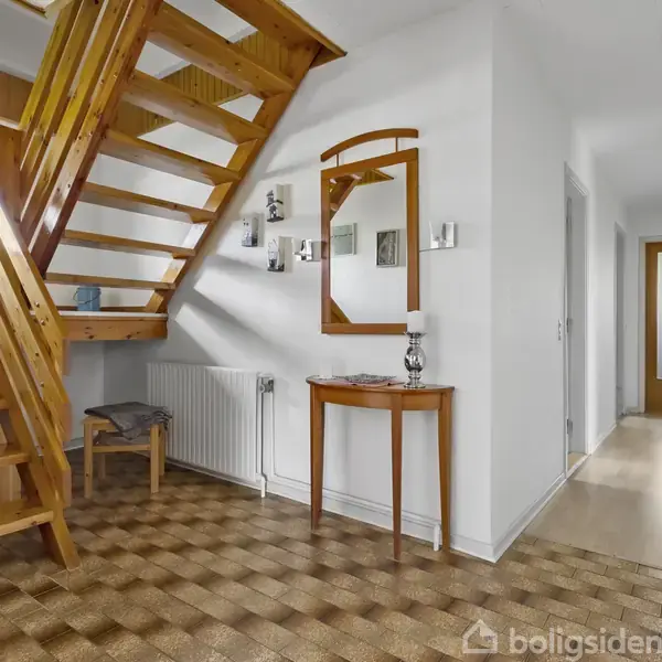 Trappe af træ fører opad i lys entre med lyst gulv, hvidmalede vægge, et lille bord og spejl. Flisegulv i forgrunden, trædør og lys i baggrunden.